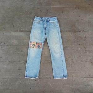 Op de afbeelding: Lichtblauwe spijkerbroek met een decoratieve patch op de linkerknie. De patch heeft een kleurrijk, geometrisch patroon in tinten rood, blauw, geel en groen. De jeans hebben een klassiek ontwerp met vijf zakken.