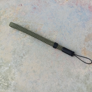 May include: A green and black patterned wrist strap with a black loop. The strap has a woven texture and is attached to a black plastic piece. The strap is lying on a textured, light gray surface.