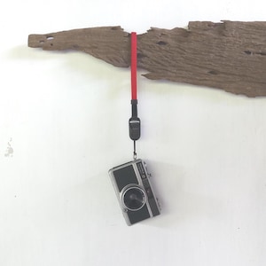 May include: A vintage silver and black camera with a red wrist strap hangs from a piece of driftwood. The camera has a lens and several buttons. The driftwood is brown and has a rough texture.