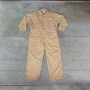 Könnte beinhalten: Beigefarbener Cord-Overall mit langen Ärmeln, einem Kragen und einem Knopfverschluss vorne. Das einteilige Kleidungsstück hat eine Brusttasche und gerade Beine. Der Overall liegt flach auf einer Holzoberfläche.