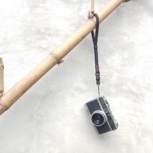 Op de afbeelding: Een vintage zwart-zilveren filmcamera met polsbandje hangt aan een bamboetak. De camera heeft een lens en een flitser. De bamboe is lichtbruin, de achtergrond lichtgrijs.