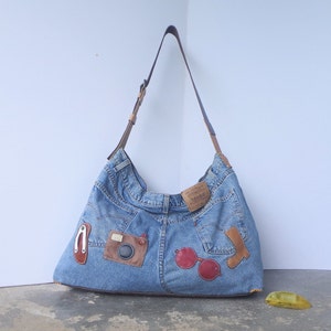 Puede incluir: Un bolso de hombro de mezclilla con detalles de cuero marrón y una correa de cuero marrón. El bolso está adornado con parches que incluyen una cámara, gafas de sol y otros elementos decorativos. El bolso está hecho de mezclilla reciclada.