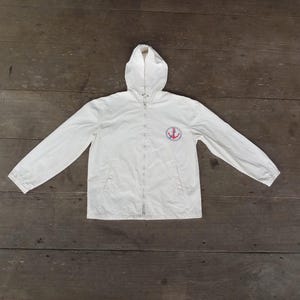 Puede incluir: Chaqueta blanca con capucha, cremallera completa y mangas largas. Un emblema de ancla rojo y azul está en el pecho izquierdo. La chaqueta está sobre una superficie de madera.