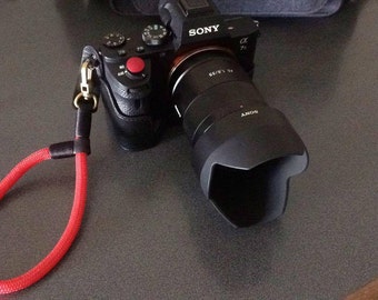 May include: A black Sony camera with a red button and a lens cap. The camera is attached to a red wrist strap.