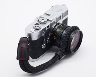 May include: A silver Leica camera with a black lens and a black braided strap with a red stitched leather end.