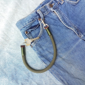 Może przedstawiać: Zielony sznurek do kluczy z przyczepionym srebrnym kluczem. Brelok wisi na kieszeni niebieskich dżinsów.