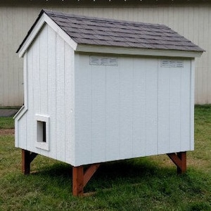 May include: White wooden chicken coop with a gray shingled roof. The coop has two small vents on the back and sits on four wooden legs.