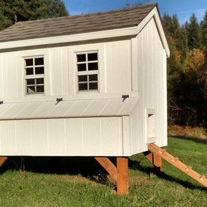 May include: White wooden chicken coop with a brown shingled roof, two windows, and a ramp for the chickens to enter. The coop is raised off the ground on wooden legs.
