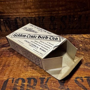 May include: An open, vintage cardboard box for "Golden Conic Herb Tea." The box is beige with black text, including "Lincoln Chemical Works, Chicago, U.S.A." and "A Remedy against Stomach Complaints." The box is on a wooden surface.