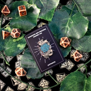May include: A blue and silver enamel pin featuring a Dungeons and Dragons dice with the text "Roll for Initiative" on a banner. The pin is on a black card with the text "Dungeons and Dragons Enamel Pin" and "@STIVAKTS". The pin is surrounded by brown and white dice.
