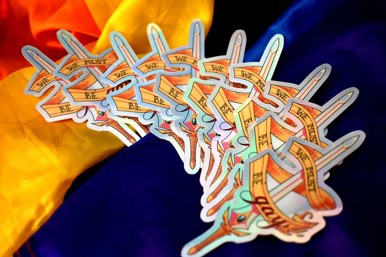May include: A stack of iridescent stickers featuring a sword and a banner with the text "We must be brave".
