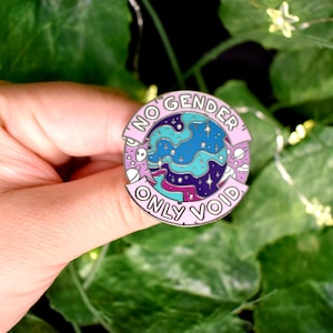 May include: A pink and blue enamel pin with a galaxy design and the text "No Gender Only Void".