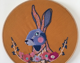 May include: An embroidered hare design in a wooden hoop. The hare is blue and pink, surrounded by colorful flowers and foliage. The background is a warm orange fabric. The text "PDF PATTERN" is at the bottom.