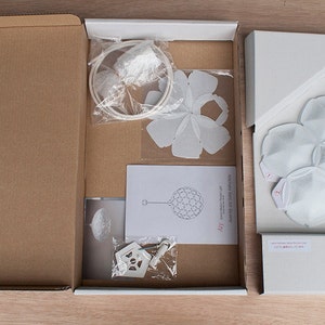 May include: A white cardboard box with three compartments. The box contains a white paper flower, a white paper sphere, and a white paper lampshade. The box is labeled "Aoi"
