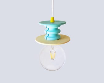 May include: A pendant light fixture featuring a white cord, a small yellow connector, a light blue tiered cylindrical component, and a flat pale yellow circular disc. A clear glass globe light bulb hangs from the center of the fixture.