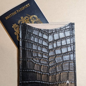 Könnte beinhalten: Schwarzer Reisepasshalter aus Kunstleder mit Krokodilprägung und einem teilweise eingelegten britischen Reisepass. Der Halter hat eine strukturierte Oberfläche und einen kleinen weißen Buchstaben "A" in der unteren rechten Ecke. Der Reisepass ist marineblau mit goldenen Buchstaben.