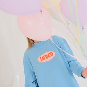 Op de afbeelding: Een lichtblauwe sweatshirt met het woord "LOVED" in een ovaal ontwerp. De persoon houdt een bos pastelkleurige ballonnen vast, waaronder roze, lavendel en geel. Lintjes in bijpassende kleuren zijn aan de ballonnen bevestigd.