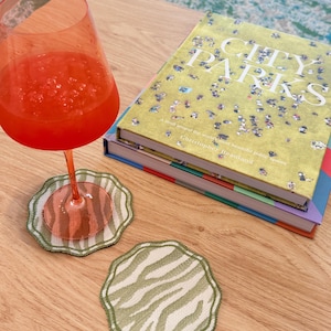 May include: Two round fabric coasters with a scalloped edge and a light green and white zebra stripe pattern. One coaster sits under a tall orange glass filled with a red beverage, while the other rests nearby on a wooden surface next to a book.
