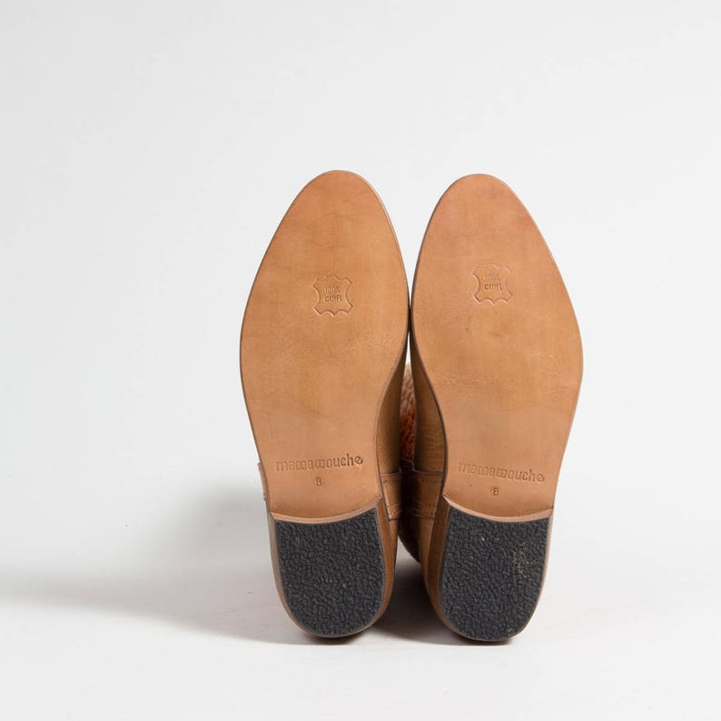 May include: A pair of tan leather boots, soles facing up, showcasing the brand name "mamamouche" and size 8. The soles are light brown leather with a black rubber heel. The boots are set against a white background.