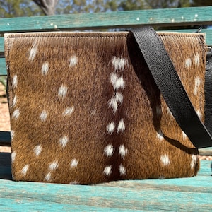 May include: A rectangular handbag with a brown and white spotted pattern, resembling deer fur. The bag has a black leather shoulder strap and a stitched top edge. The bag is sitting on a turquoise bench.