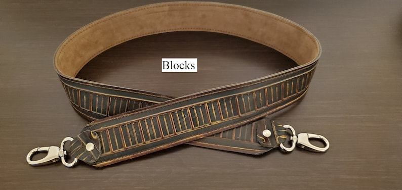 May include: A black and brown leather bag strap with a repeating rectangular design and silver-coloured clasps. The strap has a light brown interior and the word "Blocks" is in a white box.
