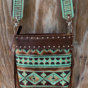 May include: A brown leather crossbody bag with a turquoise and gold geometric pattern. The bag has a fringe detail and a silver buckle closure.