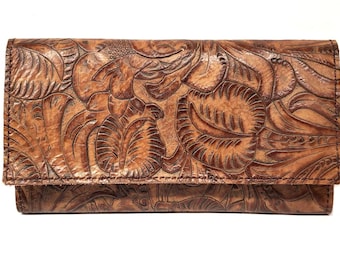 Puede incluir: Un bolso de mano de cuero marrón con un patrón floral detallado. La cartera tiene una solapa plegable y está hecha de cuero texturizado. El diseño en relieve presenta hojas y flores, creando un aspecto rústico y artesanal.
