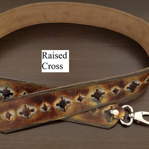 May include: A brown leather bag strap with a suede interior. The strap has a repeating pattern of cut-out crosses and silver metal clasps. The text "Raised Cross" is visible.