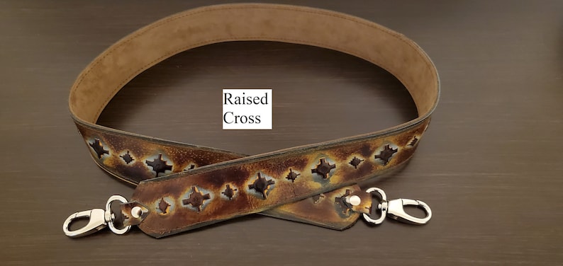May include: A brown leather bag strap with a suede interior. The strap has a repeating pattern of cut-out crosses and silver metal clasps. The text "Raised Cross" is visible.