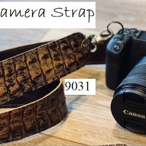 May include: A brown leather camera strap with a crocodile skin pattern is shown next to a black Canon camera with a lens. The strap has a metal clasp and the text "Leather Camera Strap" is at the top.