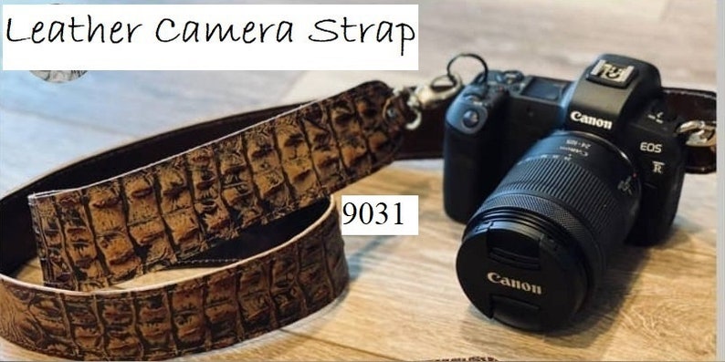 May include: A brown leather camera strap with a crocodile skin pattern is shown next to a black Canon camera with a lens. The strap has a metal clasp and the text "Leather Camera Strap" is at the top.