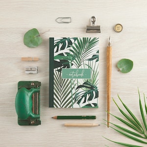 May include: A green notebook with a tropical leaf design and the word "notebook" written on the front cover. The notebook is surrounded by various stationery items, including pencils, a paper punch, a pencil sharpener, and paper clips.