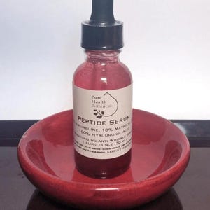 May include: A red-tinted glass bottle of Peptide Serum with a black dropper, resting in a small, round, red dish. The bottle has a white label with black text, including "Pure Health Botanicals" and "Peptide Serum". The serum is for moisturizing and anti-wrinkle.
