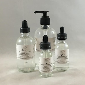 May include: Four clear glass bottles of varying sizes, filled with a liquid, are arranged on a light background. Each bottle has a black dropper or pump top and a white label with text. The labels read "Moisturizing Anti-Aging Vitamin C Serum".