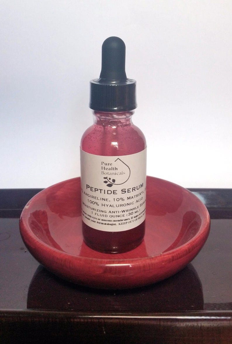 May include: A red glass bottle of Peptide Serum with a black dropper, resting on a small, red, round dish. The bottle has a white label with black text, including "Pure Health Botanicals" and "1 Fluid Ounce (30 ML)."