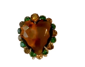 May include: A heart-shaped brooch with a large, faceted amber-colored center stone. The heart is surrounded by a halo of smaller, round gemstones in shades of green, gold, and brown. The brooch has a silver-colored metal setting.