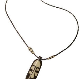 May include: A necklace featuring a pendant with a stylized face design. The pendant is cream-colored with dark brown details. The necklace has a dark chain with two striped beads and a gold-colored clasp.