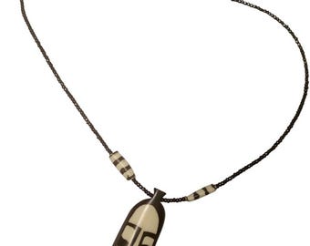 May include: A necklace featuring a pendant with a stylized face design. The pendant is cream-colored with dark brown details. The necklace has a dark chain with two striped beads and a gold-colored clasp.