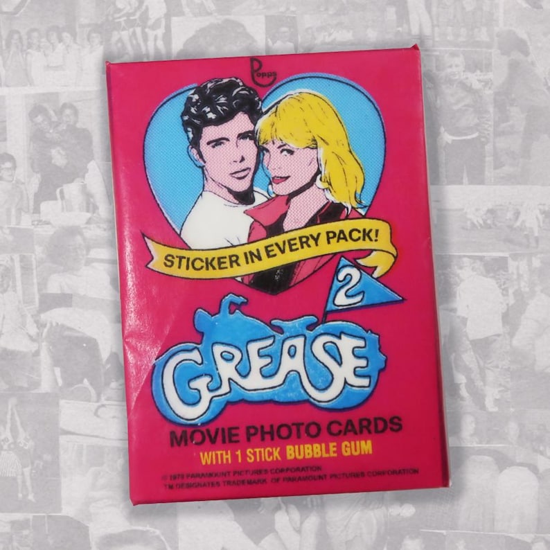Puede incluir: Una tarjeta rosa y azul con el texto "STICKER IN EVERY PACK!" y "GREASE 2" en una pancarta blanca. La tarjeta presenta una imagen de dibujos animados de Danny y Sandy de la pel&iacute;cula Grease 2. El texto "MOVIE PHOTO CARDS WITH 1 STICK BUBBLE GUM" est&aacute; en la parte inferior de la tarjeta.