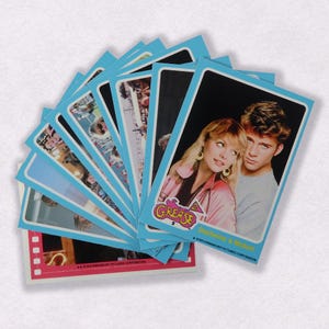 Puede incluir: Una pila de cartas coleccionables azules y rosas con im&aacute;genes de la pel&iacute;cula Grease 2. La carta superior muestra un primer plano de los actores Michelle Pfeiffer y Maxwell Caulfield. El texto de la carta dice "Grease 2" y "Michelle & Maxwell".