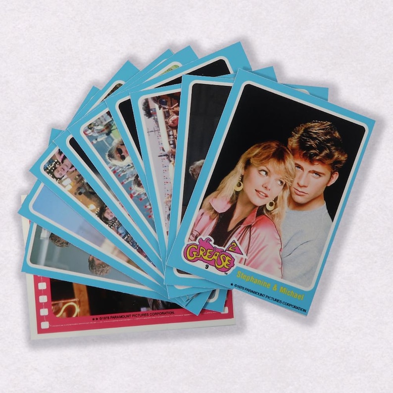 Puede incluir: Una pila de cartas coleccionables azules y rosas con im&aacute;genes de la pel&iacute;cula Grease 2. La carta superior muestra un primer plano de los actores Michelle Pfeiffer y Maxwell Caulfield. El texto de la carta dice "Grease 2" y "Michelle & Maxwell".