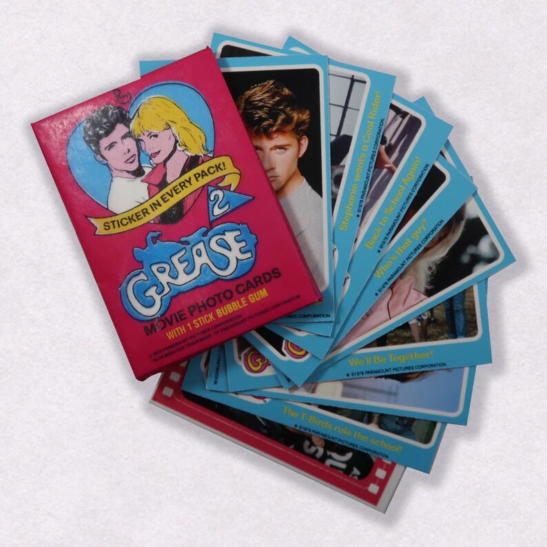 Puede incluir: Una caja rosa y azul de tarjetas de fotos de la pel&iacute;cula Grease 2 con una pegatina en cada paquete. La caja presenta una foto de los personajes principales de la pel&iacute;cula. Las tarjetas son azules con texto blanco y presentan fotos de los actores de la pel&iacute;cula.