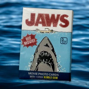 Puede incluir: Caja de tarjetas fotográficas de la película "Jaws" con chicle. La caja presenta el título de la película en rojo, una ilustración de tiburón y el texto "HIT MOVIE!" sobre un fondo azul. Se ve el logo de Topps.