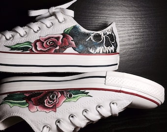 May include: A pair of white canvas shoes with hand-painted designs. One shoe features a skull and roses, while the other has a rose and green leaves.