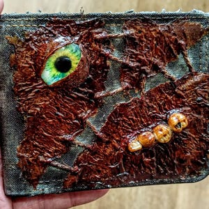 May include: A handmade wallet with a dark denim exterior. The front features a textured, brown, organic design with a large, green and yellow eye and several small, tooth-like embellishments. The wallet is held in a hand.