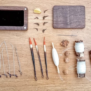 May include: Overhead view of fishing tackle laid out on a wooden surface. The image shows a wooden box, a lid, fishing floats, hooks, line, and other small components. The colour scheme is natural, with browns, whites, and reds.