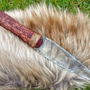 Könnte beinhalten: Ein handgefertigtes Messer mit einem Holzgriff und einer Steinklinge. Der Griff ist dunkelbraun mit einem strukturierten Muster, umwickelt mit Schnur. Die Klinge ist durchscheinend grau. Das Messer liegt auf hellbraunem Fell.