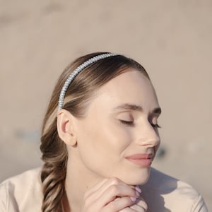 May include: A woman with long brown hair braided into a side braid wears a simple beige top and a sparkly silver headband. She has her eyes closed and is resting her chin on her hands.