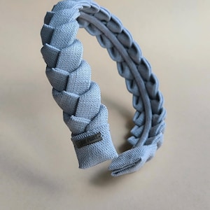 Può includere: Una fascia per capelli intrecciata azzurra. La fascia presenta un design a treccia spesso e strutturato sulla parte superiore, con una fascia più morbida e sottile sui lati. Il tessuto sembra essere lino o un materiale simile. Una piccola etichetta è attaccata all'estremità della fascia.