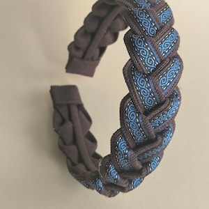 May include: Braided headband in shades of brown and blue. The headband features a woven design with a repeating pattern of blue and white swirls. The headband is made of a soft, flexible material.
